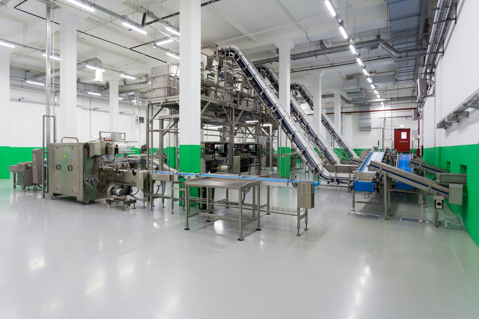 An empty food production hall: stainless conveyors angled toward a packing line, seen before a shift begins.